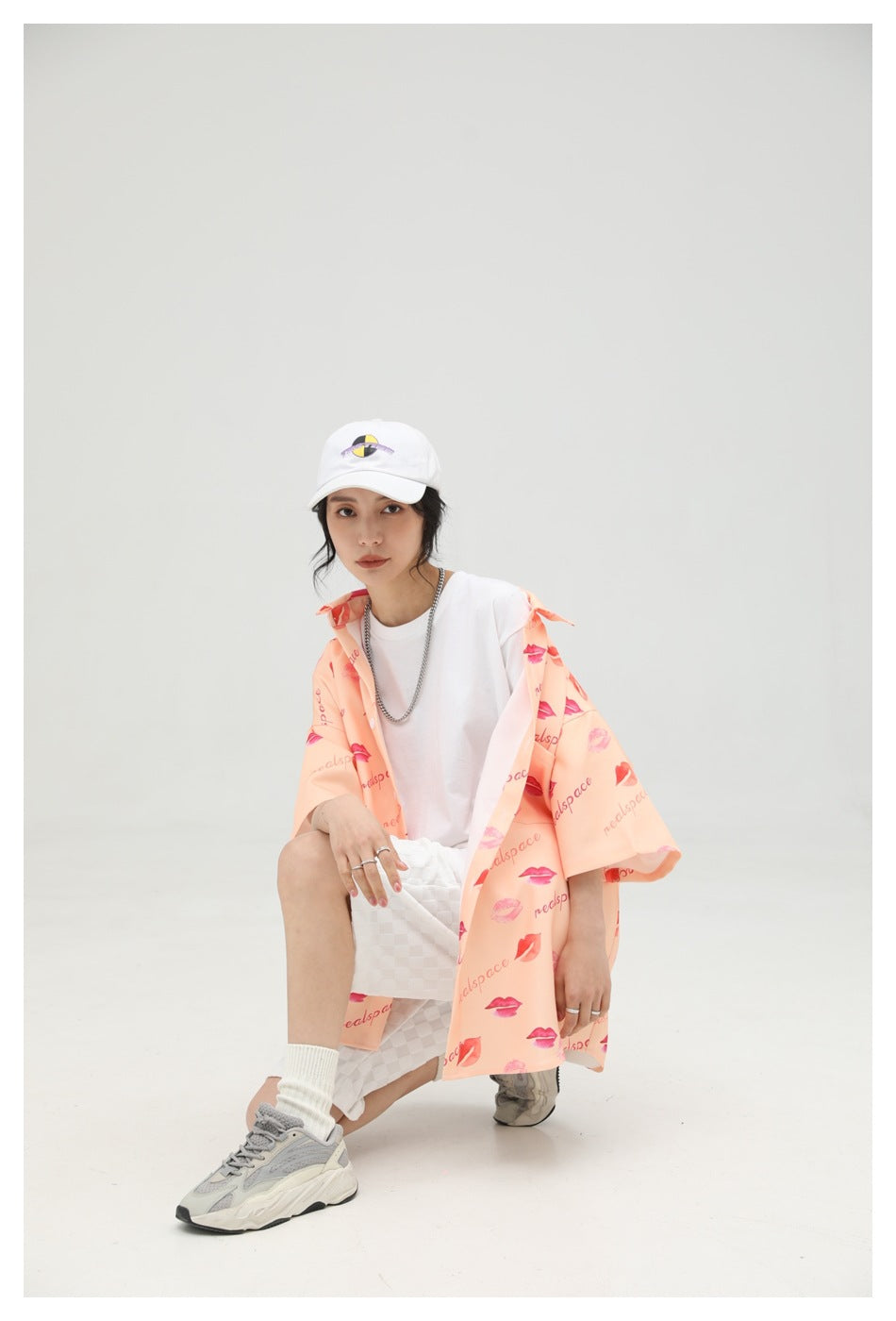 Model seated in an open peach lip-print shirt, white top, and white checkered shorts.