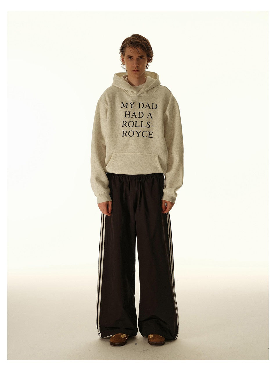 Model wearing a cream hoodie printed “MY DAD HAD A ROLLS-ROYCE” with wide black pants and side stripes.