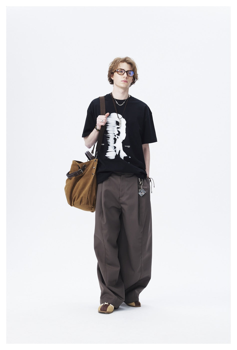 Full-length model wearing a black graphic T-shirt, loose dark trousers, glasses, and a brown shoulder bag.