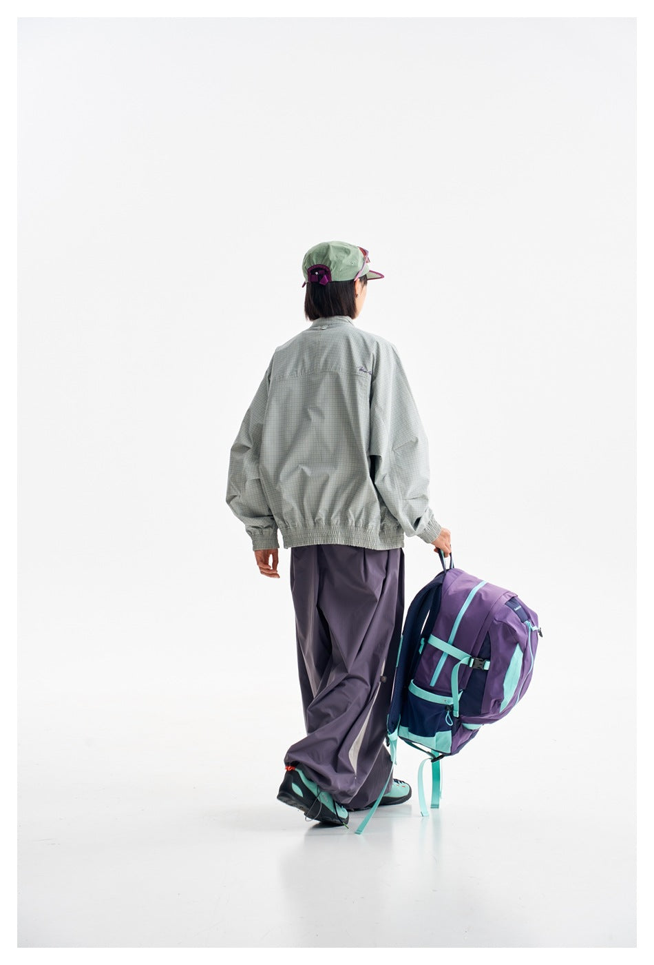 Back view of a model wearing a pale green checked jacket and loose dark purple trousers, carrying a purple backpack.