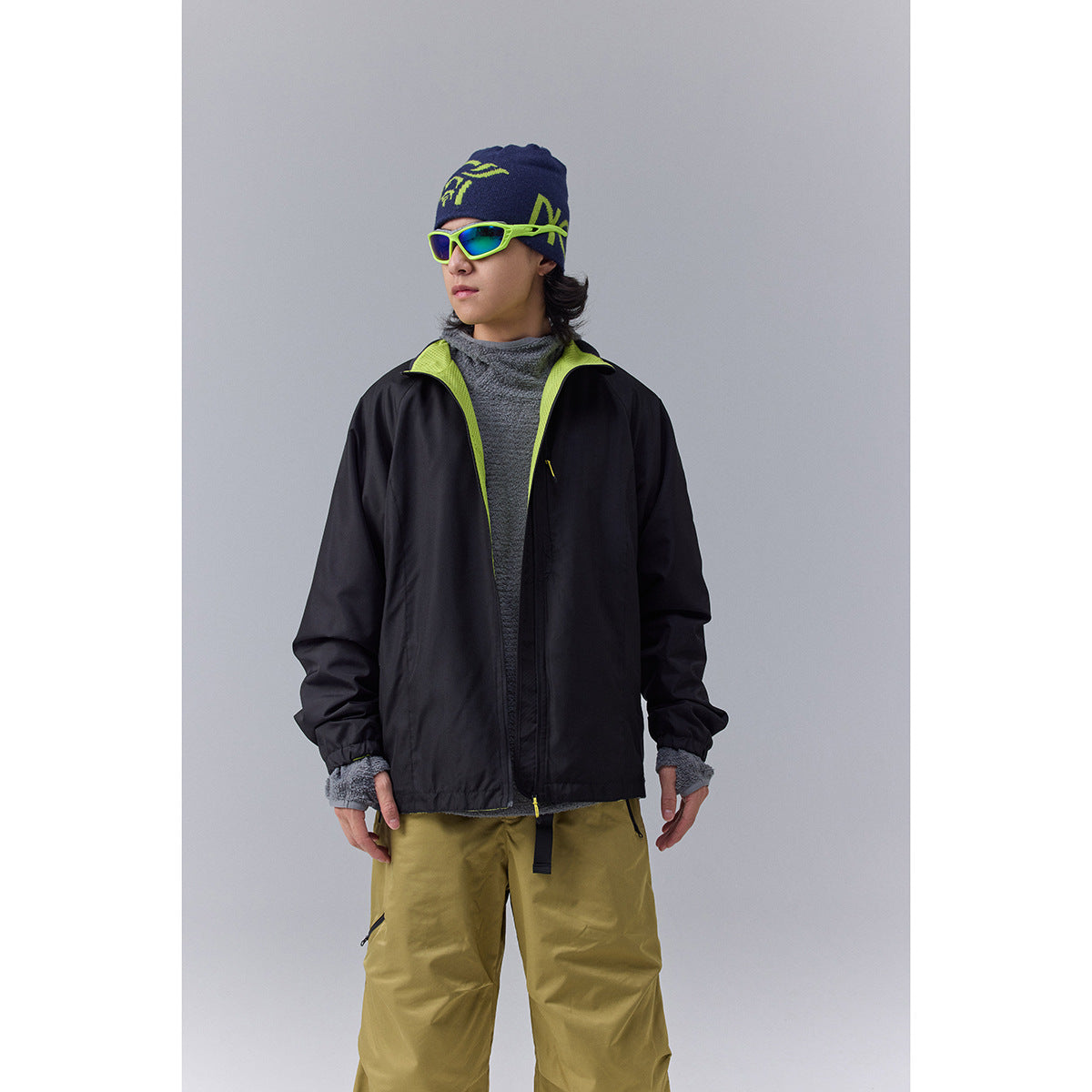 Model wearing a black zip-front jacket with lime lining, olive trousers, a navy beanie and neon sunglasses.