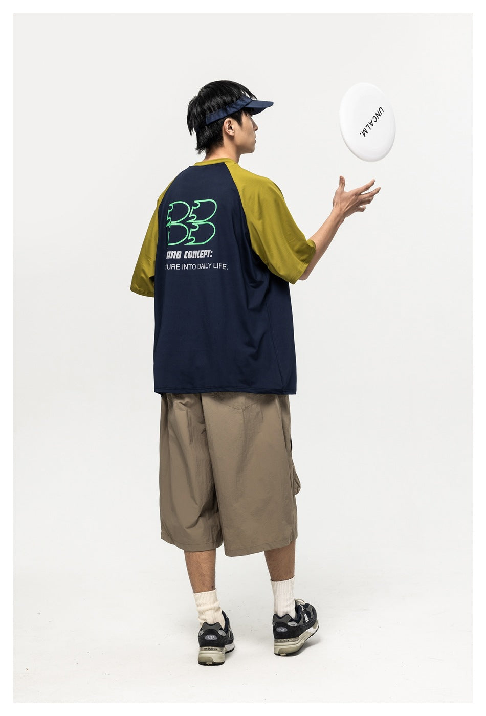 Back view of a model wearing a navy T-shirt with olive raglan sleeves and large green-and-white back graphics.