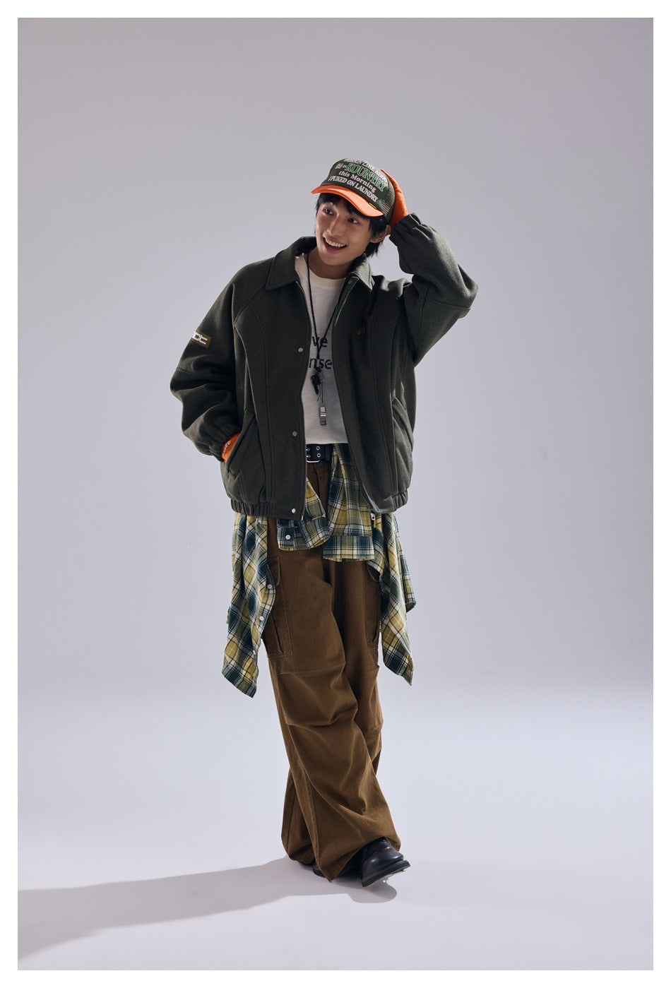 Smiling model wearing an olive green jacket with one hand in a pocket, styled with brown wide-leg pants and a cap