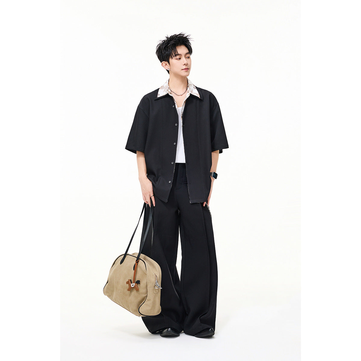 Model wearing a black short-sleeve button shirt over a floral-collar top with wide black trousers, carrying a tan bag.