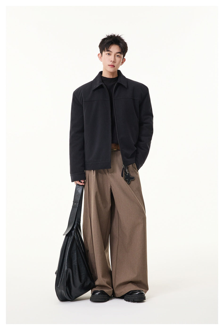 Full-length model wearing a black collared zip jacket and loose taupe trousers, carrying a black bag.