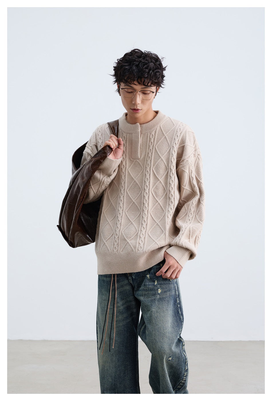 Front view of a model in a cream patterned cable-knit sweater and loose blue jeans, holding a dark tote over one shoulder.