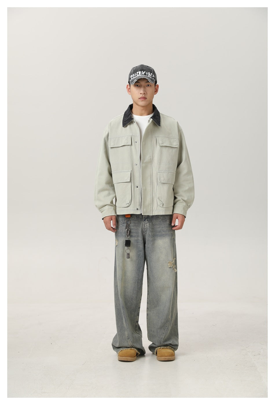 Model wearing a pale gray-green jacket with flap pockets and dark collar, with wide-leg jeans and a cap
