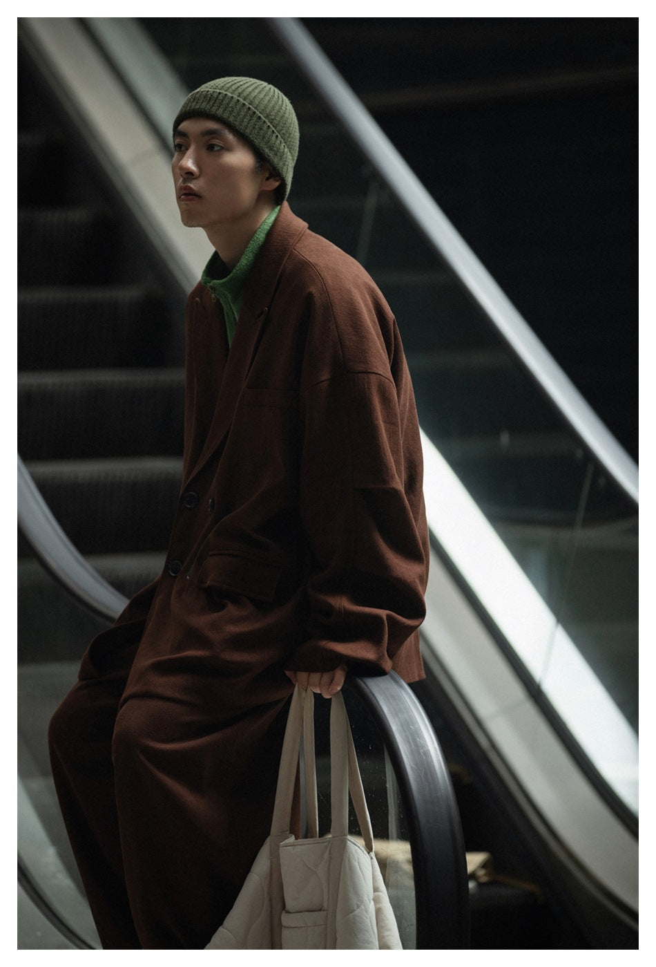 Model wearing a brown oversized coat, green zip-neck top, and olive beanie, seated beside an escalator.