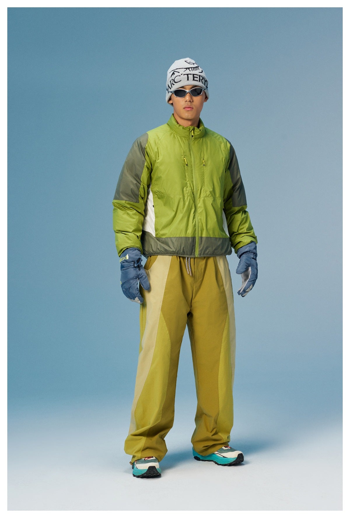 Full-length view of a man wearing a lime-green jacket with gray panels, mustard trousers, blue gloves, sunglasses, and a
