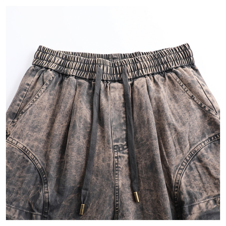 Front view of brown-gray patterned pants with a gathered waistband, black drawstrings, angled pockets, and curved panel