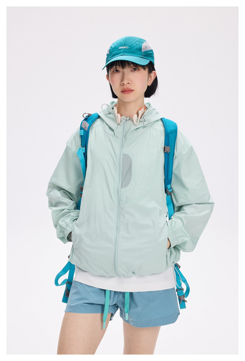 Model wearing a pale mint hooded zip jacket with blue shorts, turquoise cap, and bright blue backpack straps.