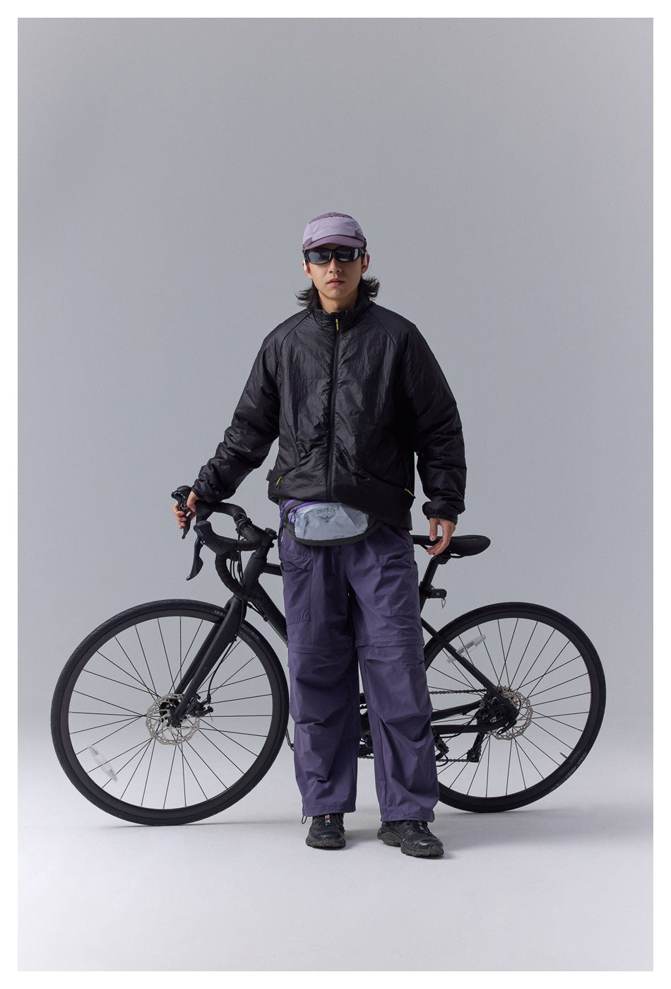 Model wears a black zip-front jacket and loose purple cargo pants beside a road bike.