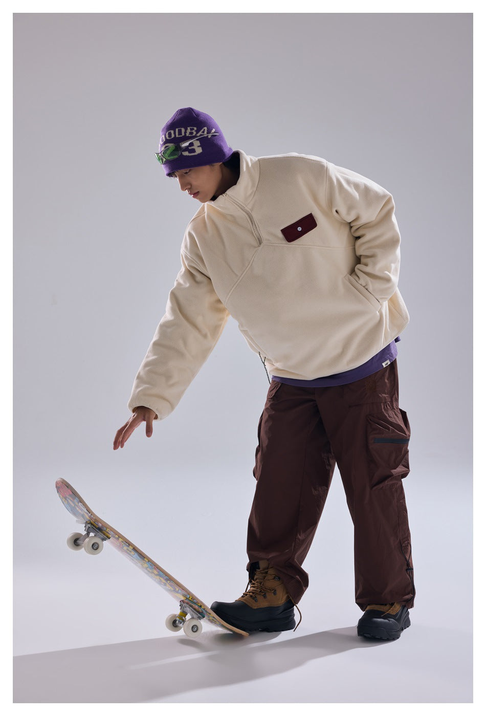 Model wearing an ivory quarter-zip pullover and brown cargo pants, reaching toward a skateboard.