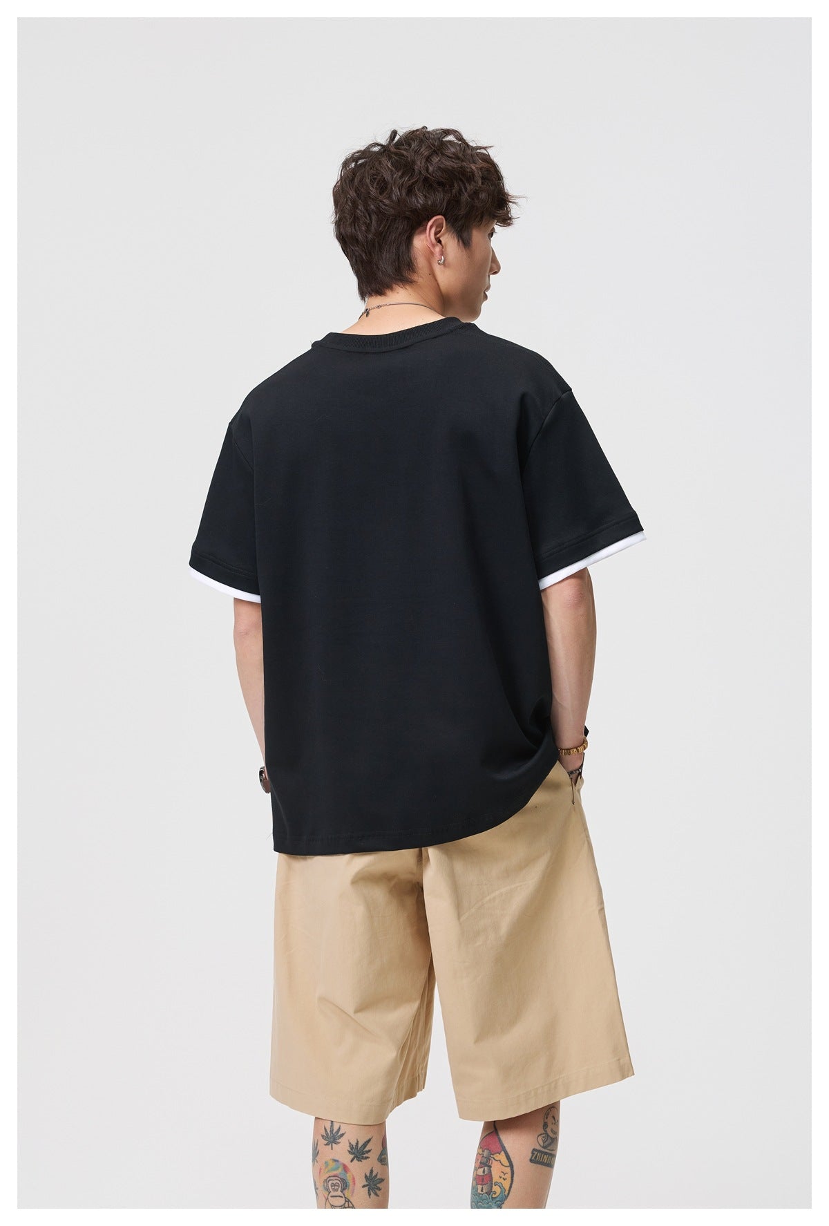 Back view of a model wearing a black oversized T-shirt with white sleeve layers and beige shorts.