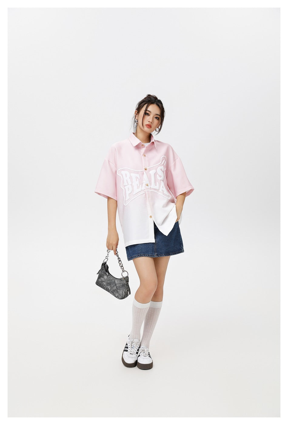 Model wearing an oversized pink-to-white button-up shirt with raised white lettering and a dark denim skirt.