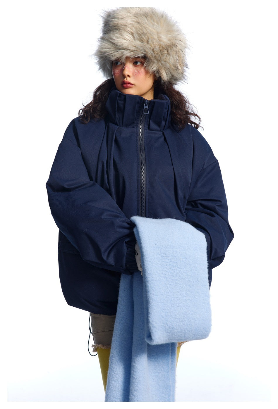 Model wearing a navy high-collar zip jacket and holding a pale blue scarf, with a fur hat and beige shorts.