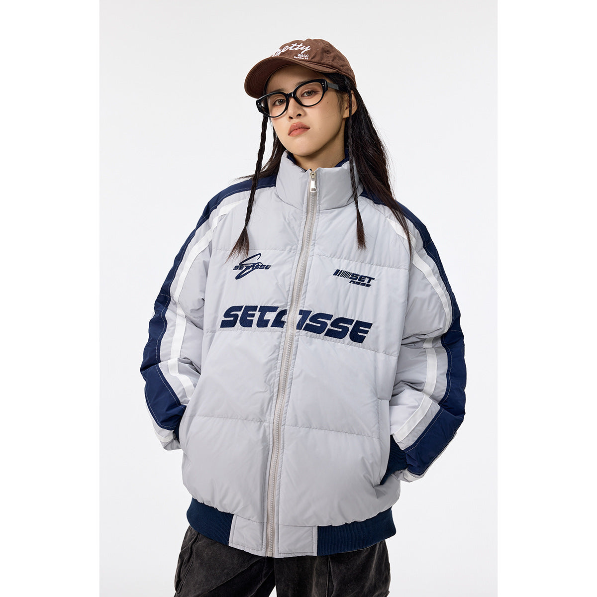 Model wearing a light gray padded zip jacket with navy panels, white sleeve stripes and dark chest lettering.