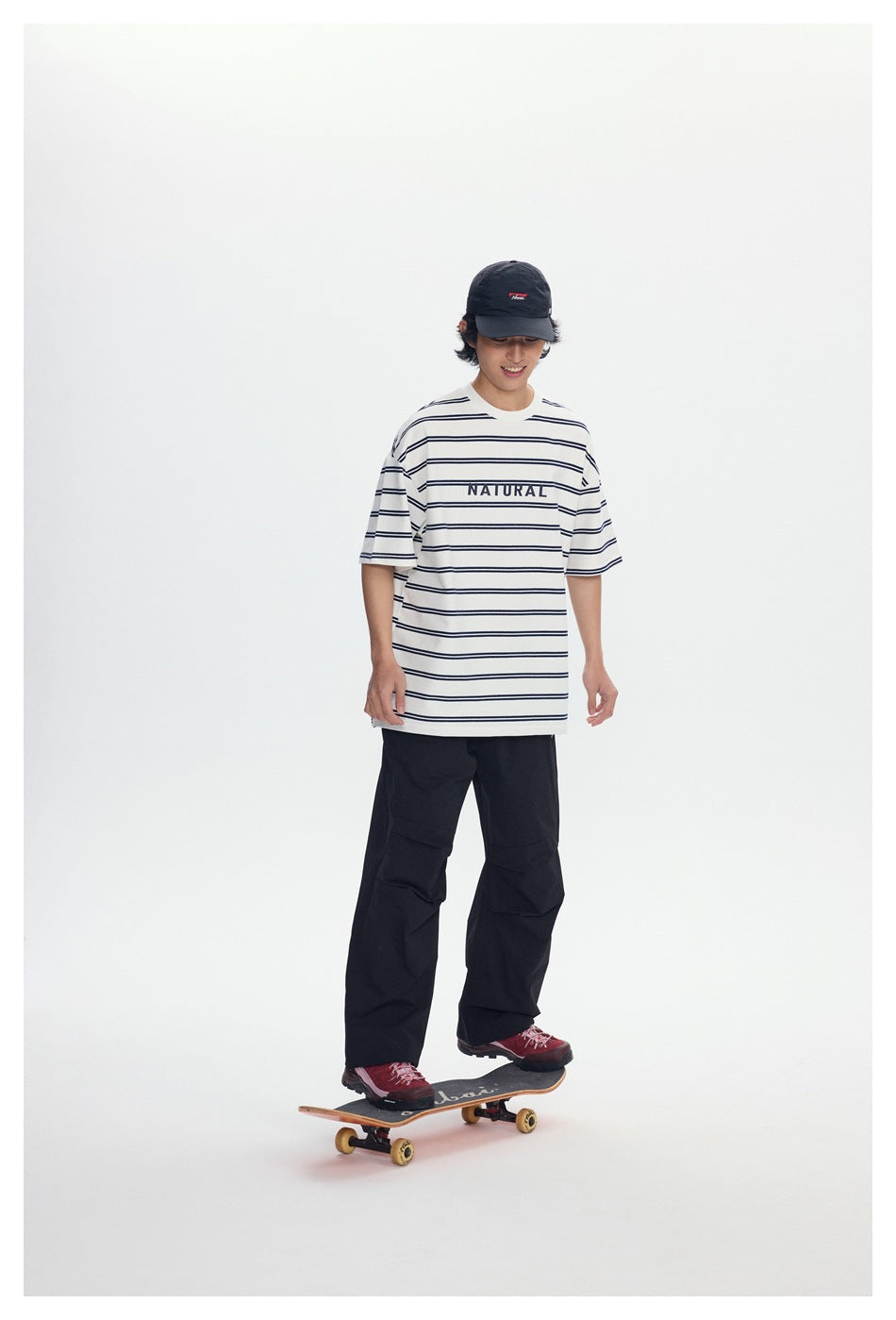 Model wearing a white T-shirt with thin navy stripes and NATURAL text, loose black pants, and a black cap while standing on a