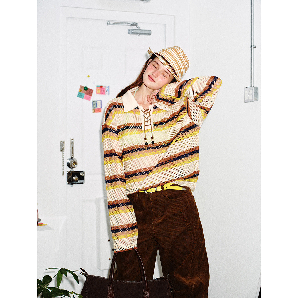 Model wearing a cream open-knit sweater with yellow, brown, and navy stripes, a lace-up collar, and a striped hat.