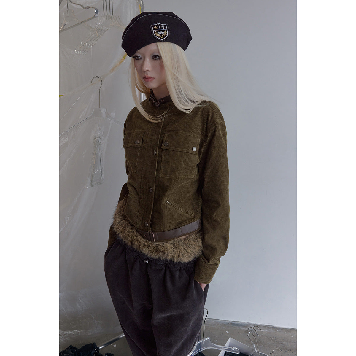 Model wearing an olive jacket with snap pockets, a brown belt and fur-trimmed hem, paired with dark pants and a black beret.