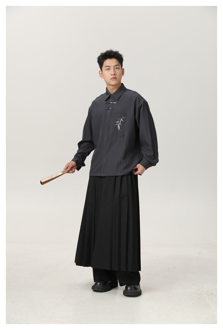 Full-length front view of a man wearing a charcoal shirt with metal front fasteners and bamboo chest embroidery, paired with