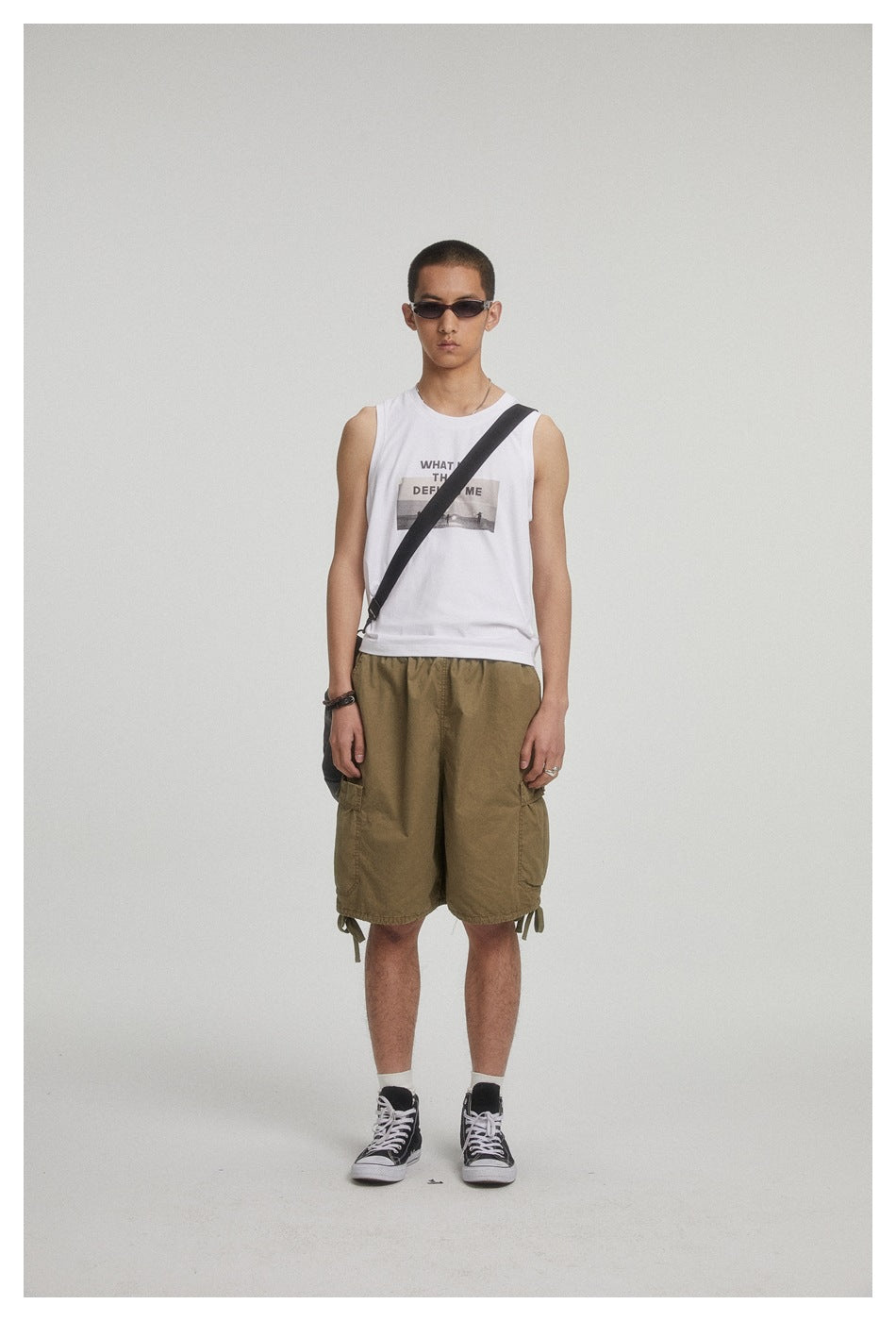 Full-length model in a white graphic sleeveless shirt and olive cargo shorts, with sunglasses and a crossbody bag.