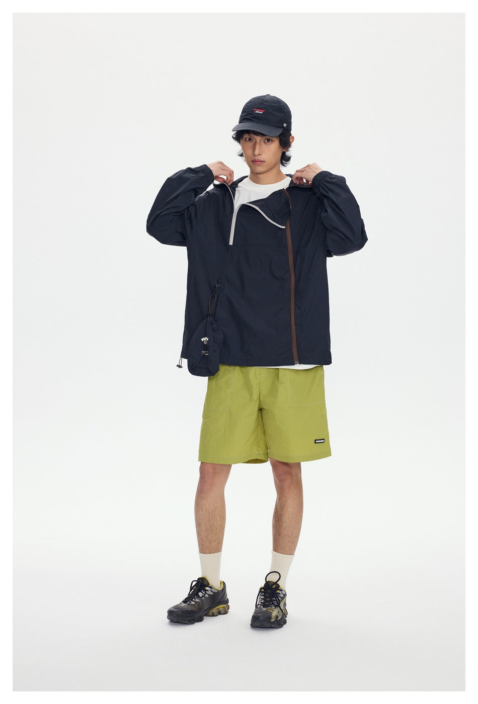 Front view of a model wearing a dark hooded pullover jacket with contrasting zips and lime-green shorts.