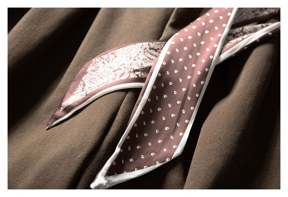 A dusty rose narrow scarf with white edging and small white motifs lies over brown fabric.