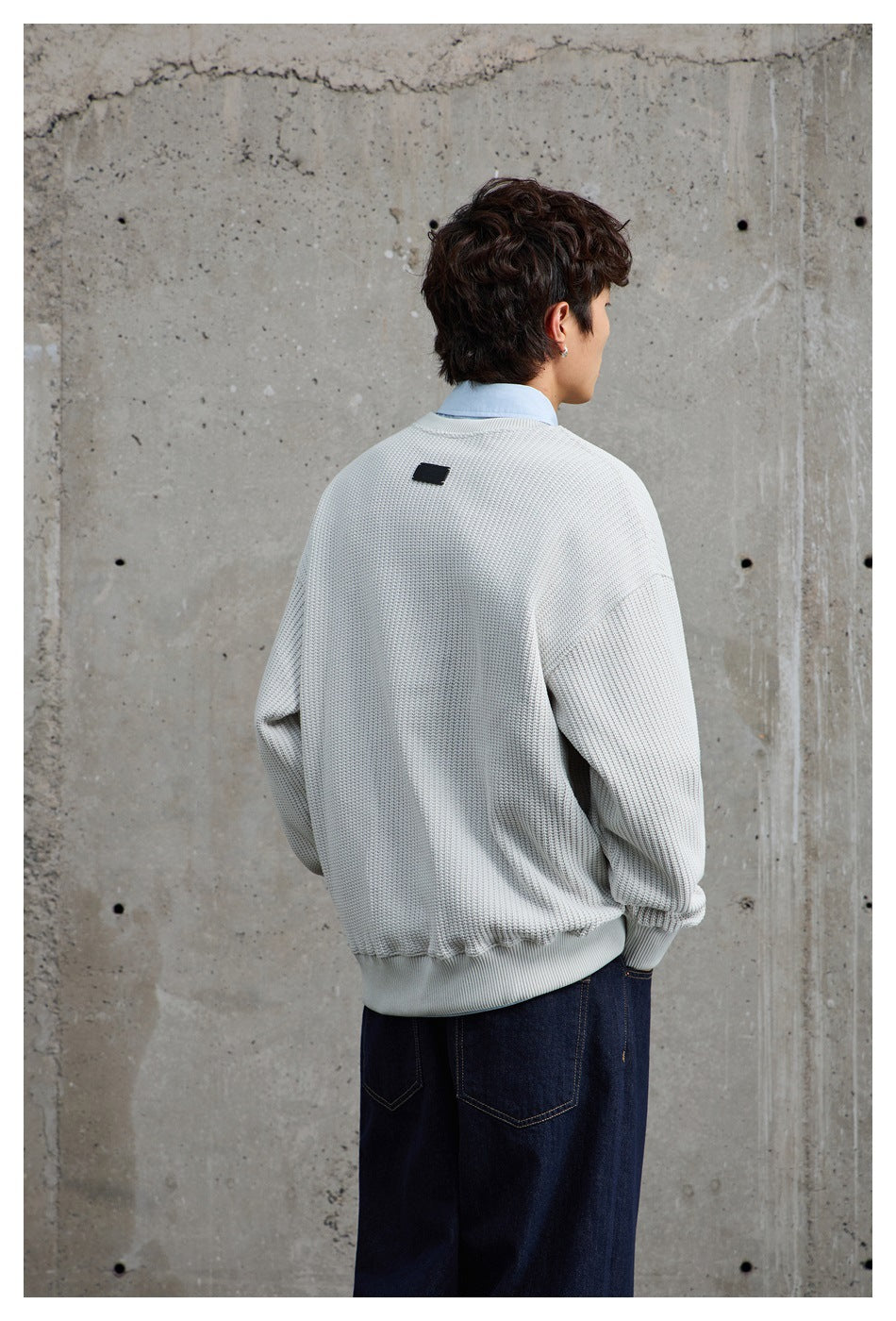 Back view of a model wearing a light gray textured crewneck sweater with ribbed trim and dark jeans.