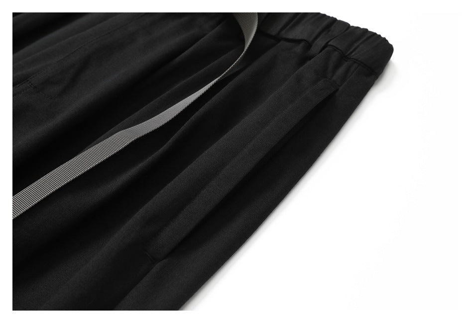 Close-up of black trousers showing the elastic waistband, belt, and stitched side pocket.