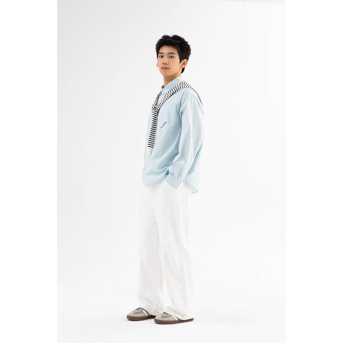 Full-length model wearing a pale blue button-up shirt, striped sweater over the shoulders, white trousers, and sneakers.