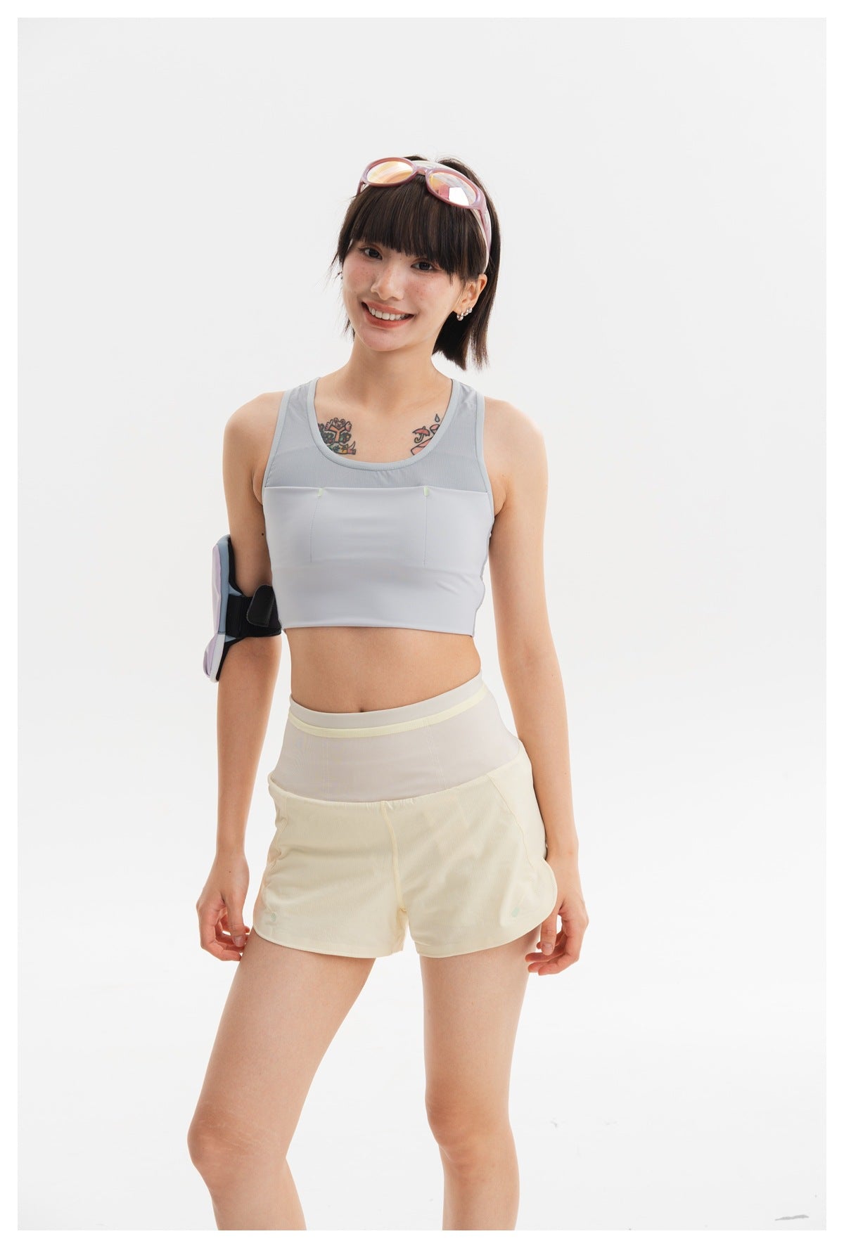 Model wearing a light gray cropped tank top and pale yellow shorts, with a padded armband on a white studio background.