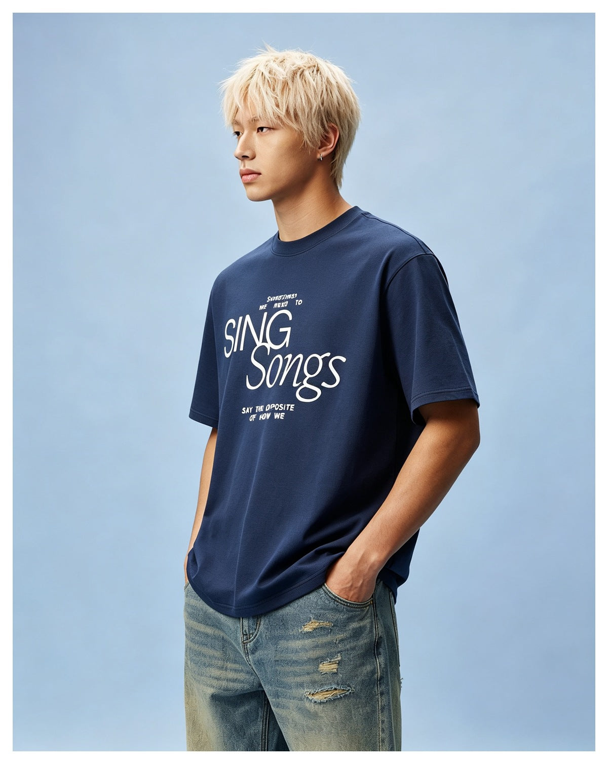Man with blond hair wearing a navy blue 'SING Songs' graphic T-shirt with distressed light-wash jeans on a blue background