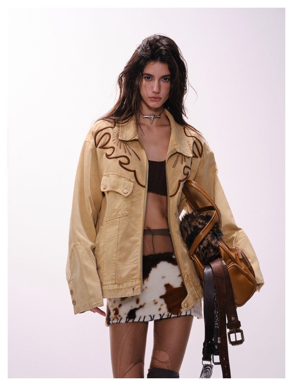 Model wearing a tan oversized jacket with dark swirling embroidery and flap pockets over a patterned mini skirt.