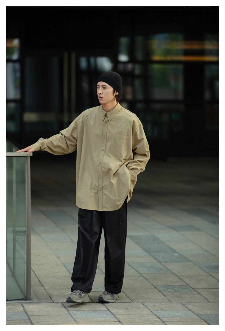 Model wearing a loose khaki button-up shirt with a chest pocket and black trousers.