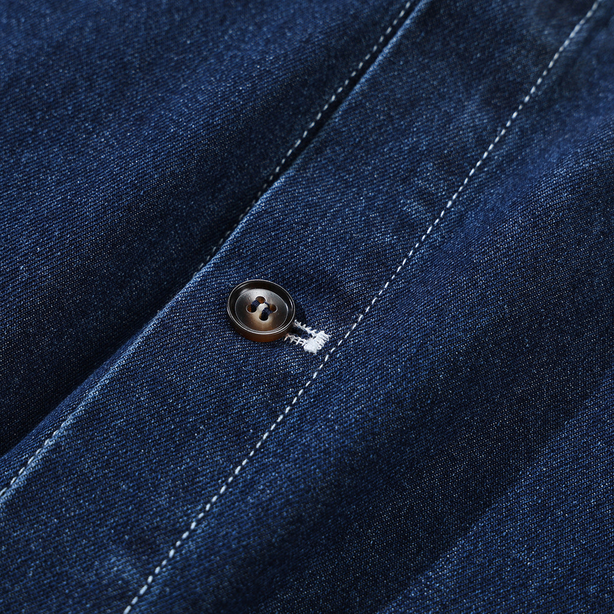 Close-up of a dark button and white stitching on the placket of a dark indigo denim shirt.