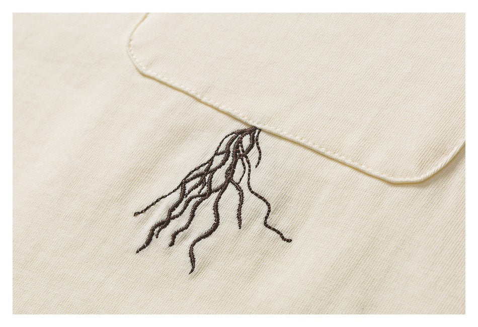 Close-up of a cream garment pocket edge with dark embroidered tree roots extending below it.