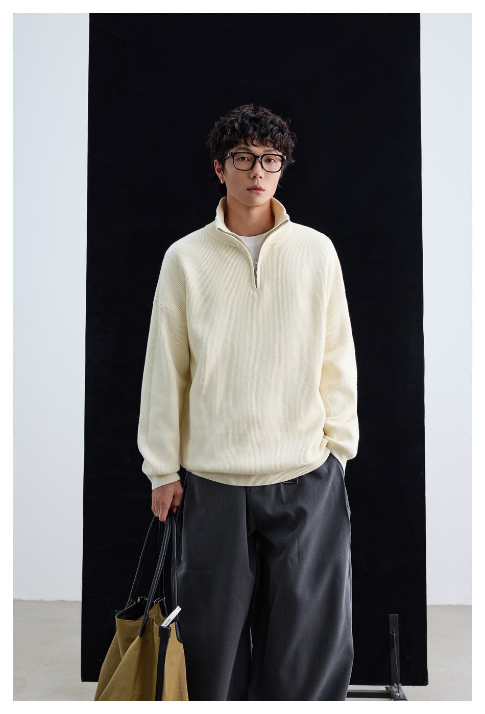 Model wearing cream quarter-zip knit sweater with gray wide trousers, holding a tote bag against a black backdrop
