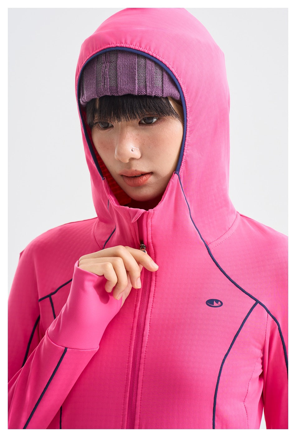 Close-up of a pink jacket's hood, purple knit cap, front zipper, navy piping, and small chest emblem.