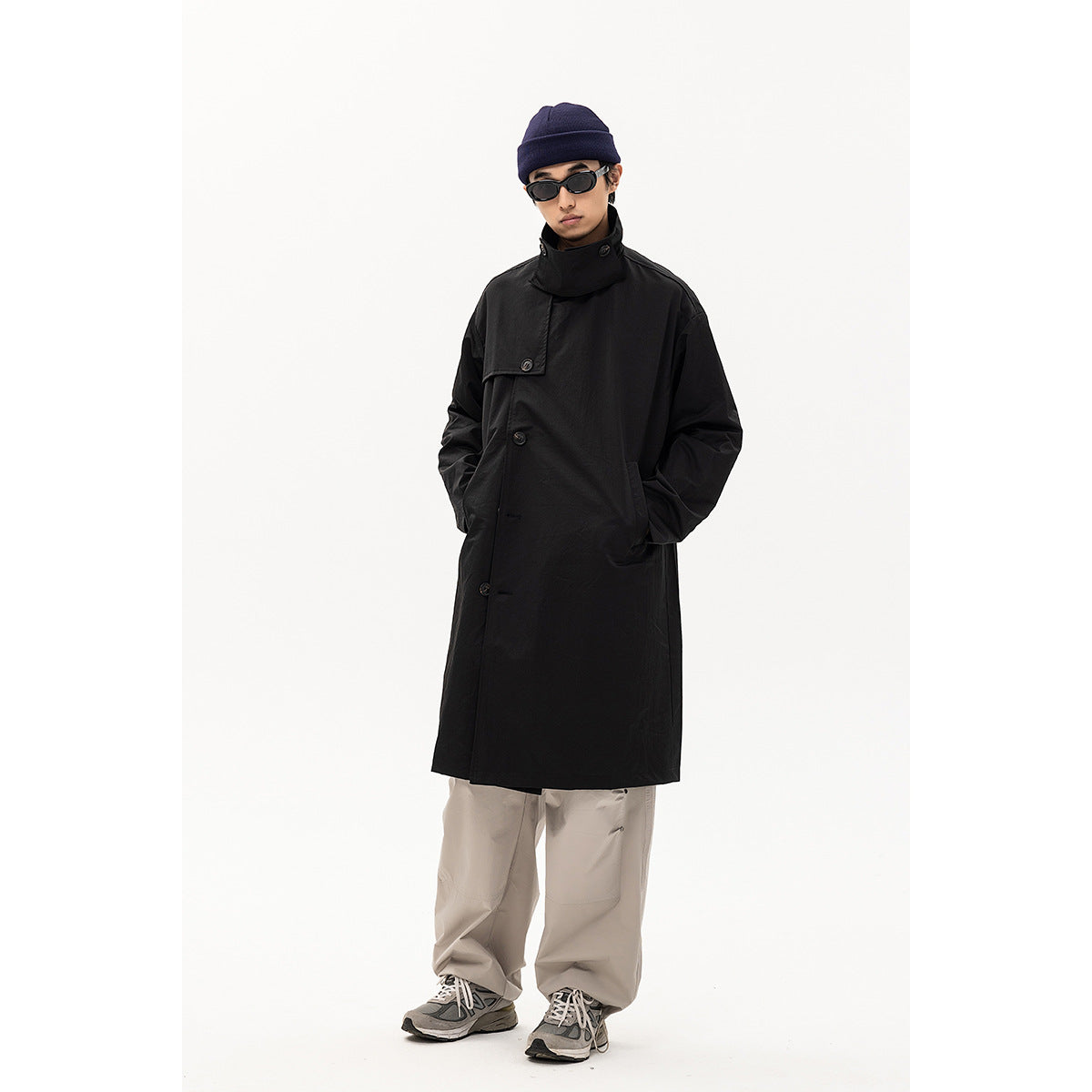 Full-length front view of a model in a long black button-front coat, navy beanie and loose gray trousers.