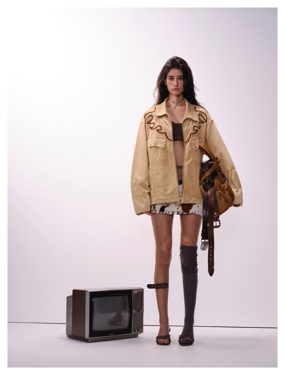 Model wearing an oversized sand-brown embroidered western jacket open over a brown top and cow-print skirt