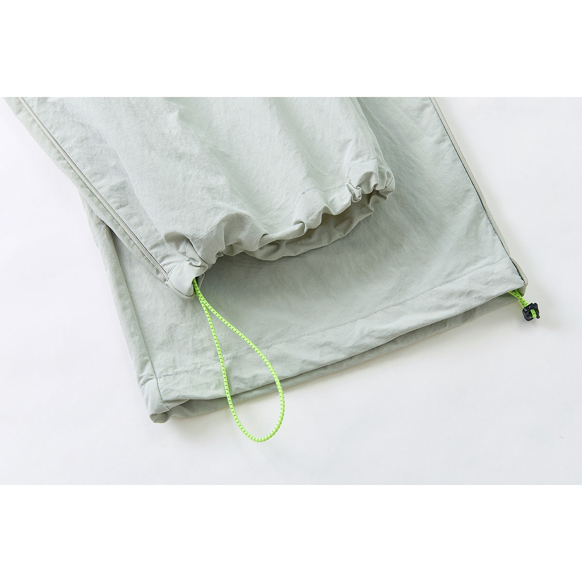 Close-up of pale sage pants hems, showing a gathered inner cuff and bright lime adjustable drawcords.