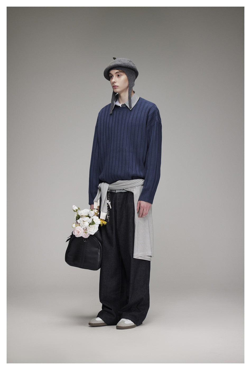 Model wearing a blue ribbed sweater over a checked shirt, with dark wide-leg jeans and a gray hat.