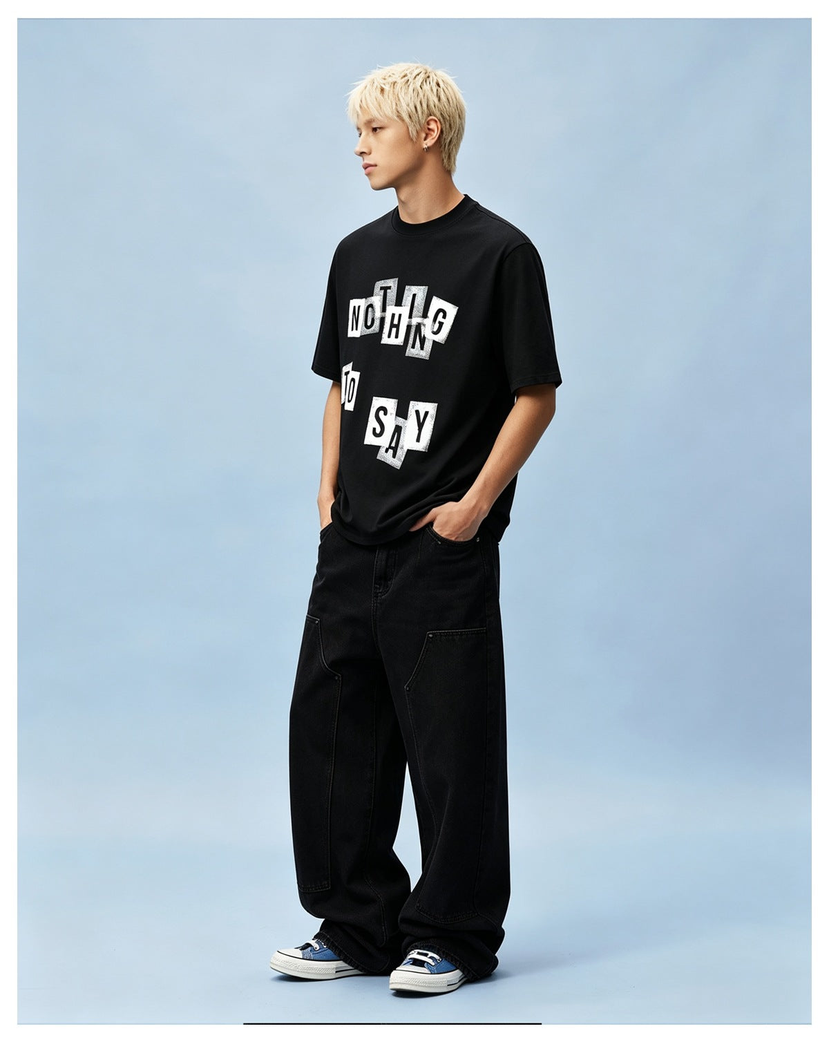 Model wearing black graphic crew-neck T-shirt with wide-leg black jeans and blue sneakers against a light blue backdrop