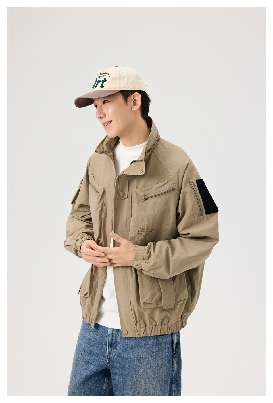 Man wearing a tan utility jacket with a high collar, zip chest pockets, black sleeve patch, and elastic cuffs.