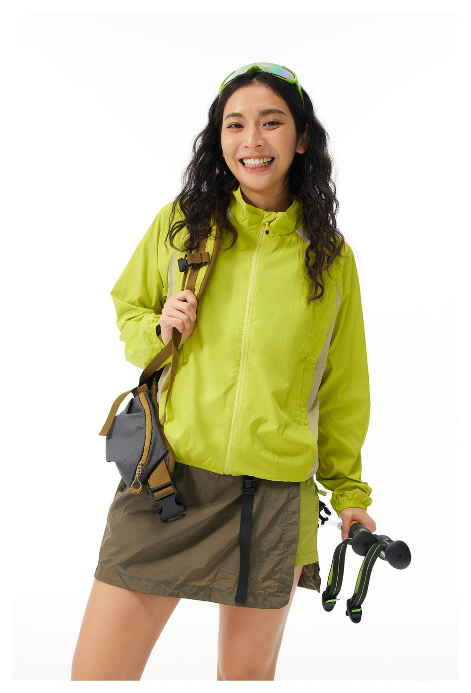 Smiling woman wearing a yellow-green zip jacket with a backpack strap, hip bag and trekking pole