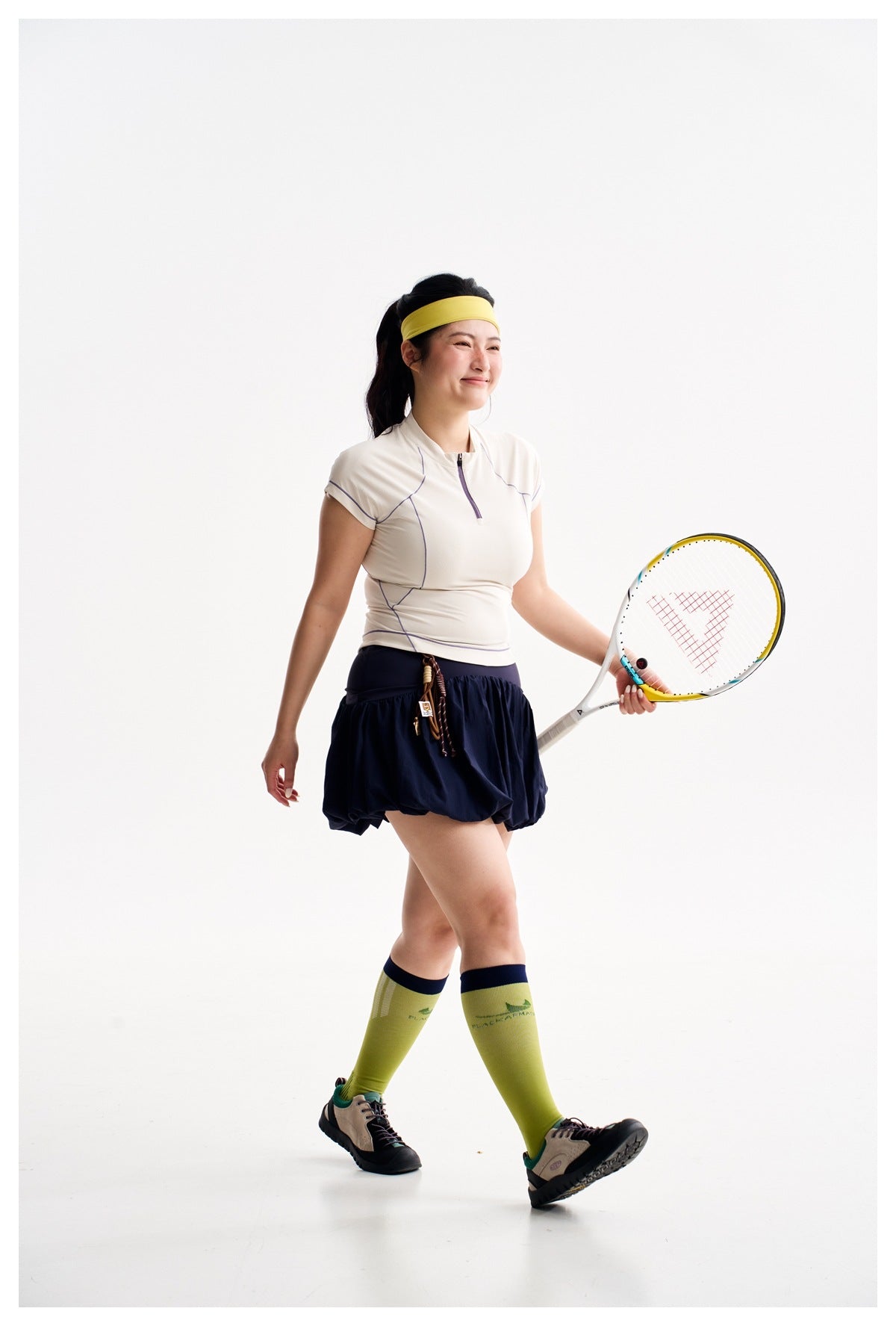 Model holding a tennis racket, wearing an ivory zip-neck top, navy bubble skirt, and lime knee-high socks.