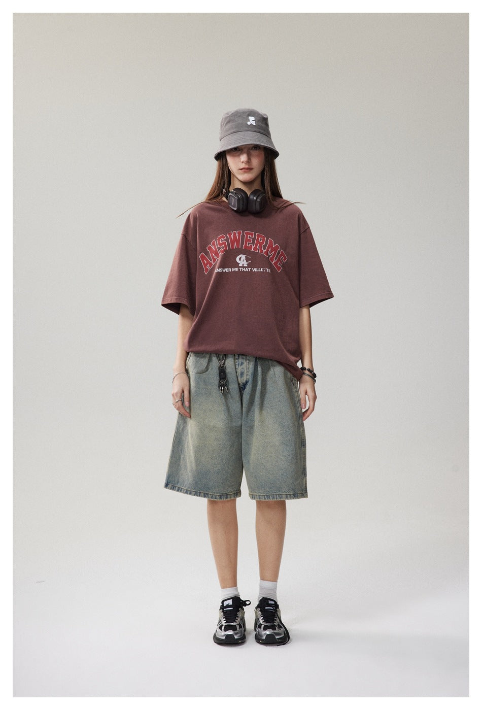 Full-length model wearing a burgundy graphic T-shirt, gray bucket hat, and loose light-wash denim shorts.