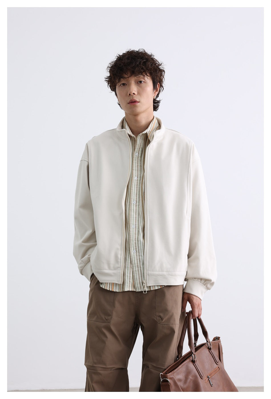 Front view of a model wearing an open off-white zip jacket over a striped shirt with taupe trousers.