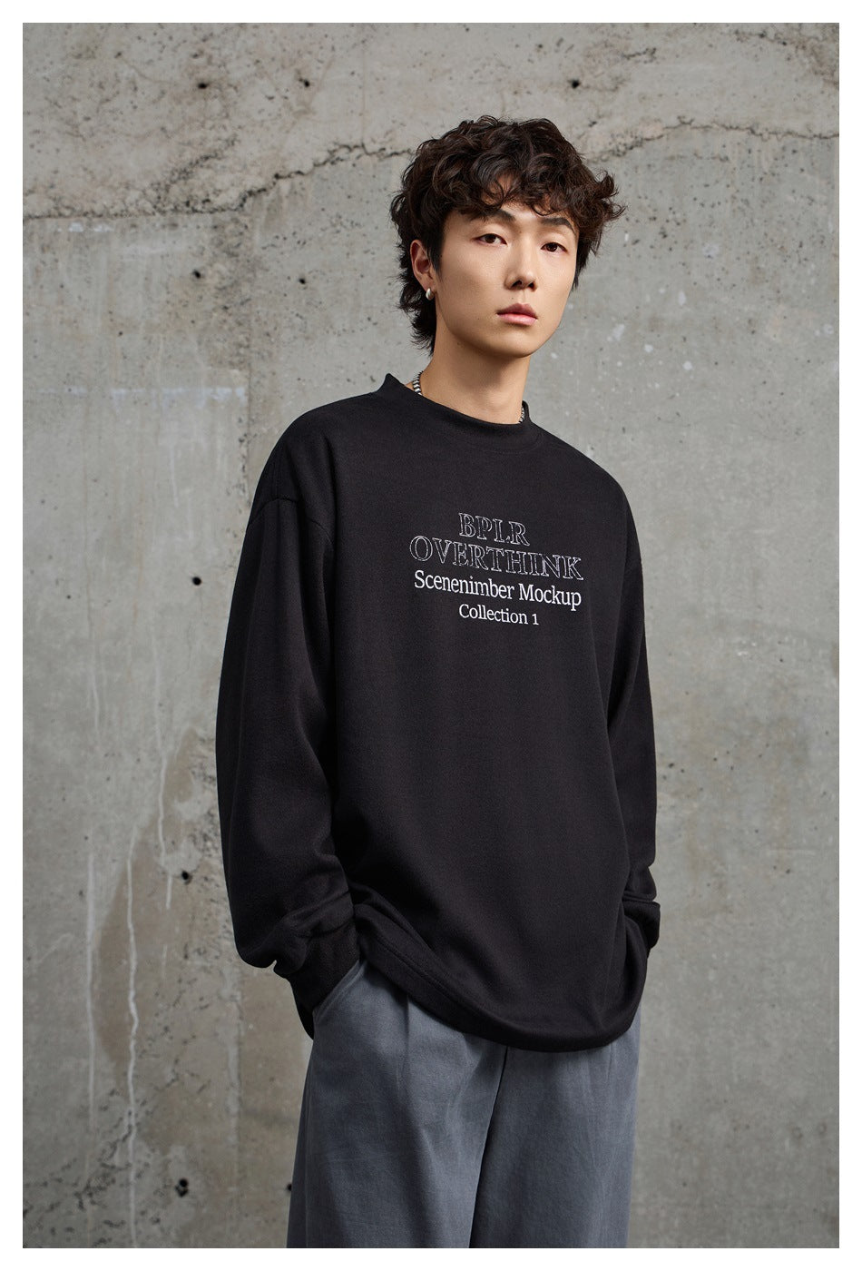 Model wearing an oversized black long-sleeve crewneck with white text on the chest and loose gray trousers.